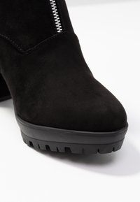 Botín negro de ante con puntera redondeada, cierre lateral de cremallera y suela de goma texturizada que presenta un patrón de relieve para mayor agarre.