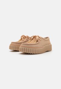 Chaussures en suede beige avec un bout rond, une semelle en caoutchouc proéminente et des stries texturées. Dotées de lacets et d'accents de couture orange contrastants.