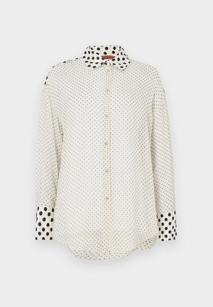 Λευκό πουκάμισο με κουμπιά και μακριά μανίκια, με μικρές μαύρες πουά σε όλο το ύφασμα και μεγαλύτερες μαύρες πουά στον γιακά και στις μανσέτες.