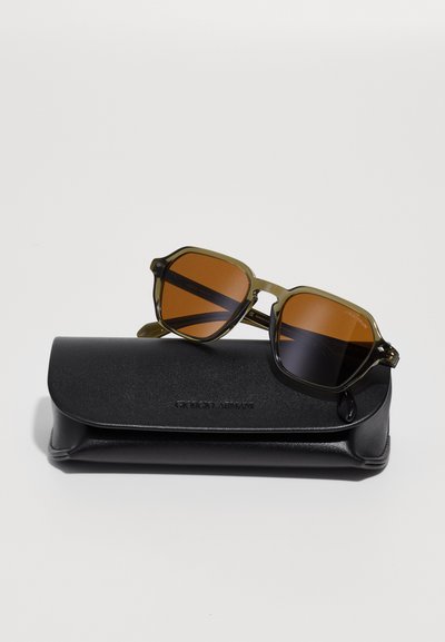 Olive green sunglasses with brown lenses, featuring an octagonal frame. Accompanied by a black hard case with embossed logo.