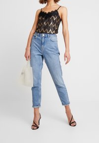 Černý a zlatý top s chevronovým vzorem a krajkovým lemem, spárovaný se světle modrými džíny s vysokým pasem a černými páskovými podpatky. Drží bílou síťovanou tašku.