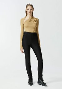 Bež majica s dugim rukavima i naboranom tkaninom, uparena s crnim visokim uskim hlačama i crnim čizmama sa šiljastim vrhom i metalnim detaljima.