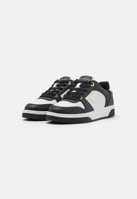 Schwarz-weiße Sneaker mit einem perforierten Design, flachen Schnürsenkeln, goldenen Akzenten und Gummisohle, mit einem Logo auf der Zunge.