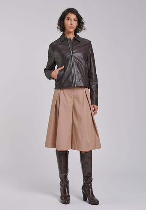 Kobieta w ciemnobrązowej skórzanej kurtce, beżowej spódnicy midi oraz dopasowanych brązowych kozakach za kolano, stojąca na tle jasnoszarego tła.