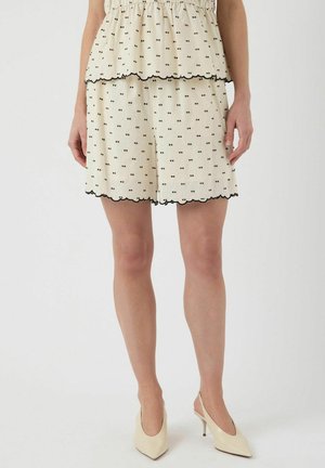 Krämfärgade shorts med svart dekorativt mönster och scalloped fåll, tillverkade av lättviktigt tyg; matchas med ljusa klackar.
