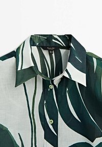 Chemise à col boutonnée avec grands motifs abstraits de feuilles vertes et blanches et deux boutons visibles sur le devant.