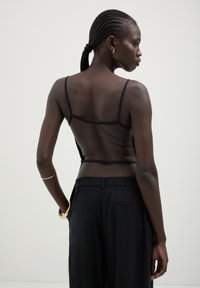 Top nero trasparente con spalline sottili, abbinato a pantaloni neri a vita alta. La modella ha i capelli intrecciati e indossa un bracciale d'oro.