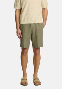 Oluļu zaļi lina šorti ar brīvu piegriezumu, kas aprīkoti ar priekšējām kabatām un gludu, teksturētu virsmu. Apvienoti ar vieglu, teksturētu krējuma krāsas krekla.