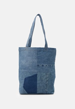 Blaue Patchwork-Jeans-Tasche mit zwei Schulterriemen und sichtbarer Tasche unten links vor einfachem Hintergrund.
