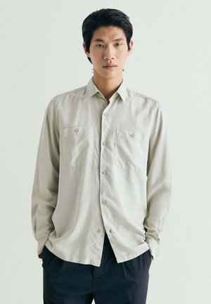 Joven de pie con las manos en los bolsillos, vistiendo una camisa beige clara abotonada y pantalones oscuros contra un fondo claro y sencillo.