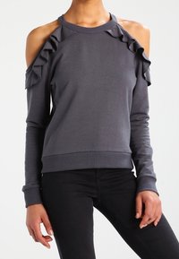 Sudadera gris con diseño de hombros al aire y acentos con volantes. Mangas largas y puños acanalados. Tela suave, forma ajustada y estilo relajado.