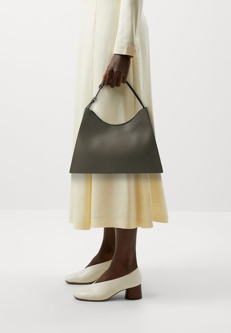 Sac en cuir gris avec un dessus arrondi, tenu par une main portant une longue robe de couleur claire, associé à des chaussures à talons blocs blanches.