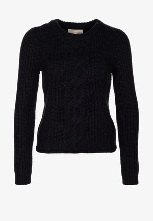 Femme marchant vêtue d'un pull noir à torsades, d'un pantalon skinny noir, de chaussures noires brillantes à lacets, et portant un sac à bandoulière gris à franges.