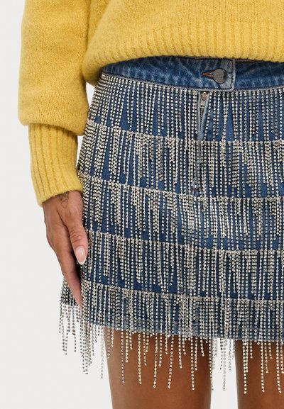 Jeansrock mit silberner Perlenfransenverzierung, ausgestattet mit einem Reißverschluss und blauer Textur, kombiniert mit einem gelben Strickpullover.