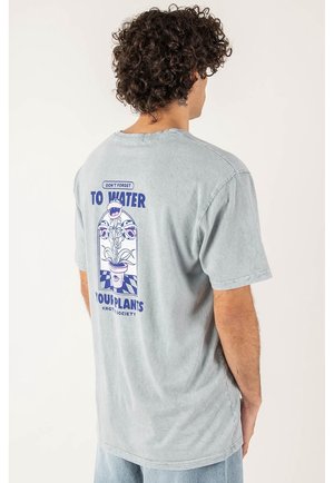 Man met krullend haar in een vervaagde grijze T-shirt met een blauwe plantgrafiek en de tekst "Vergeet niet je planten water te geven" op de achterkant.