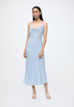 Femme debout dans une robe midi bleu clair sans manches à fines bretelles et sandales blanches, sur un fond blanc uni.