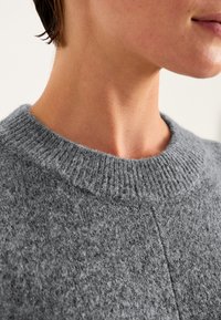 Harmaa neulepaita, jossa on korkea, ribattu kaulus ja pehmeä, tekstuuri pintakuvio; sisältää hienovaraisia saumarakenteita.