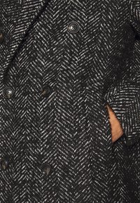 Černobílý kabát s dvouřadým zapínáním a vzorem rybí kostí, výraznými knoflíky a texturovanou tkaninou, s bočními kapsami.
