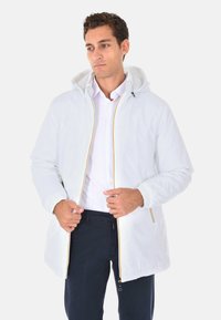Giacca bianca leggera con cappuccio, caratterizzata da dettagli gialli sulla zip e polsini elasticizzati, indossata sopra una maglietta bianca e pantaloni scuri.