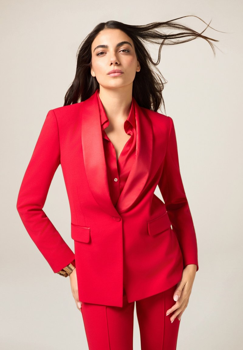 Czerwony garnitur na miarę z satynowym kołnierzem w stylu szalowym, długimi rękawami i dwiema kieszeniami. Koszula również czerwona, o gładkiej fakturze i detalach z guzikami.