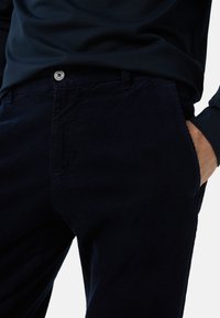 Marineblå kordrembukser med en struktureret overflade, der har en sølvknap og sidelommer. Stofmetoden har lodrette ribbede mønstre.