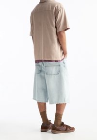 Camisa de cuello corta en color beige con un acento morado; pantalones cortos azul claro hasta la rodilla; zapatillas marrones y calcetines oliva.