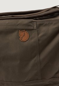 Saco de tecido verde oliva escuro com textura suave, apresentando um emblema redondo de couro castanho com um design estilizado de animal.