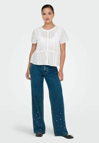 Weißes Kurzarmoberteil mit Durchbrüchen und Knoten in der Taille, kombiniert mit blauen, weit geschnittenen Jeans mit Perlenverzierungen.