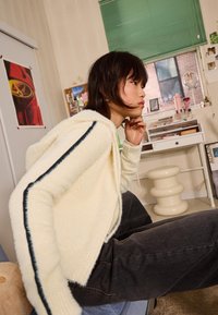 Kremna puhovka z navadno črto po rokavu, nosena čez temne kavbojke; v ozadju je plakat in pisalna miza.