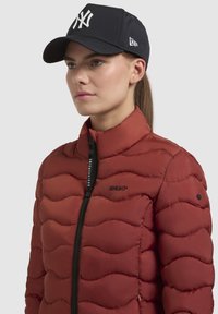 Rode pufferjas met een golvenpatroon, ritsluiting en zwart logo. Draag het met een marineblauwe baseballcap met een wit logo en vizier.