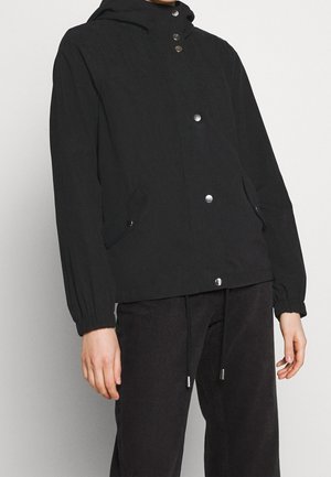 Personne portant une veste à capuche noire avec des boutons-pression argentés, des poignets élastiques et deux poches à rabat à l'avant, associée à un pantalon foncé.