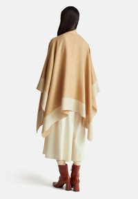 Beige pončo z vlněné směsi s krémovými akcenty, s volným tvarem a vrstveným designem. Nalezla se přes bílé sukně a červené kožené boty na podpatku.