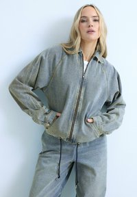 Blondynka ubrana w szarą, oversize'ową kurtkę dżinsową na zamek i pasujące luźne spodnie dżinsowe, stojąca z rękami w kieszeniach kurtki.