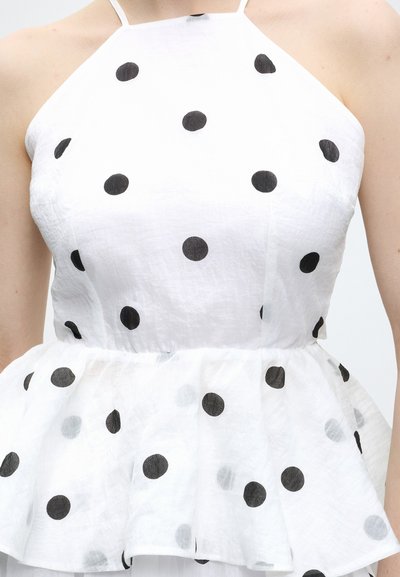 Haut sans manches blanc à pois noirs avec taille peplum porté par une personne à la peau claire.