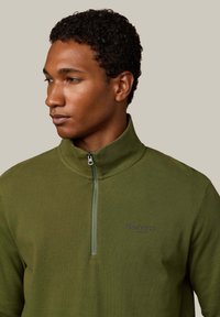 Sudadera verde con cremallera y cuello alto, de tejido texturizado, con un logo bordado en azul marino. Presenta una cremallera frontal y un diseño ajustado.