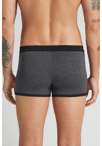 Zadní pohled na osobu nosící přiléhavé tmavě šedé pruhované boxerky s černým pasem a lemováním nohavic, zobrazující tetování na pažích a horní části zad.