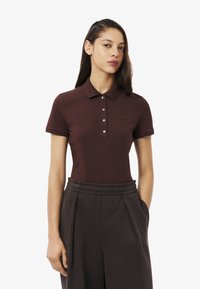 Polo bordeaux avec un col classique, patte de boutonnage à deux boutons et un logo discret près de la poitrine. Porté avec un pantalon sombre, coupe relax.