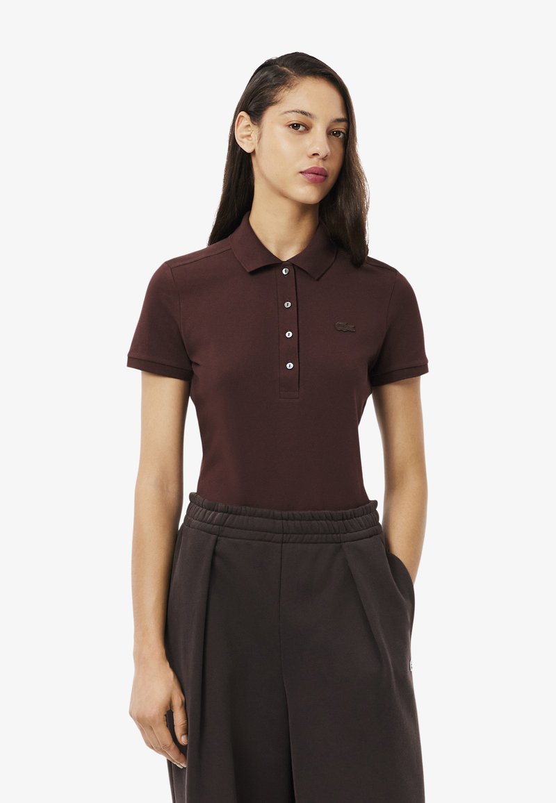 Polo bordeaux avec un col classique, patte de boutonnage à deux boutons et un logo discret près de la poitrine. Porté avec un pantalon sombre, coupe relax.