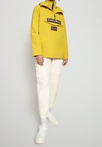 Gele waterdichte jas met een halve rits, grote voorkantzak en logodetails. Gecombineerd met witte cargobroek en witte sneakers.