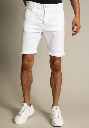 Man draagt opgerolde witte denim shorts en witte sneakers, staand tegen een effen beige achtergrond.