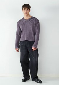 Lila gerippter Strickpullover mit langen Ärmeln, V-Ausschnitt und rohem Saum, kombiniert mit locker sitzenden schwarzen Jeans und glänzenden schwarzen Schuhen.