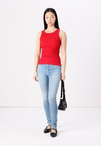 Rød ribbet tanktop, blå stramme jeans, sorte nitter flade sko, lille sort håndtaske. Model står mod en neutral baggrund.