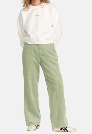 Kvinde iført hvid sweatshirt med "RVCA"-logo, lysegrønne bredbenede bukser i fløjl og hvide sneakers med sorte striber.