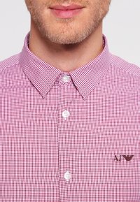 Muž se světlým plnovousem, oblečený v červené a bílé kostkované košili s knoflíky a špičatým límcem, na které je vyšité logo AJ.