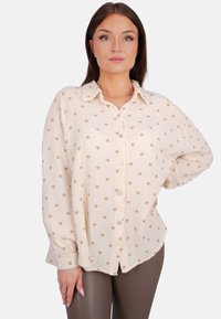 Cremfarbiges Hemd mit strukturiertem Stoff, das mit einem Muster aus kleinen braunen Herzen versehen ist, sowie langen Ärmeln mit Knopfmanschetten.