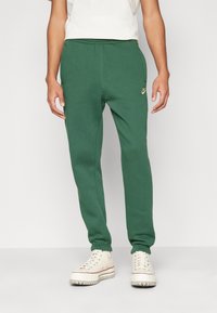 Zelené tepláky ze soft materiálu, s vyšitým zlatým logem Nike, elastickým pasem a zúženými manžetami. Noseny s bílými teniskami.