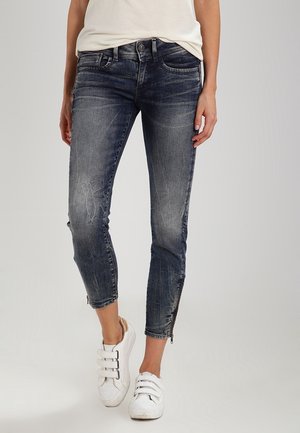 Personne portant un jean skinny bleu foncé délavé avec des fermetures éclair aux chevilles et des baskets blanches à scratch, debout devant un fond uni.