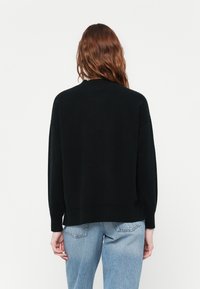 Personne avec de longs cheveux roux ondulés portant un pull noir à manches longues et un jean bleu clair, de dos contre un fond uni.