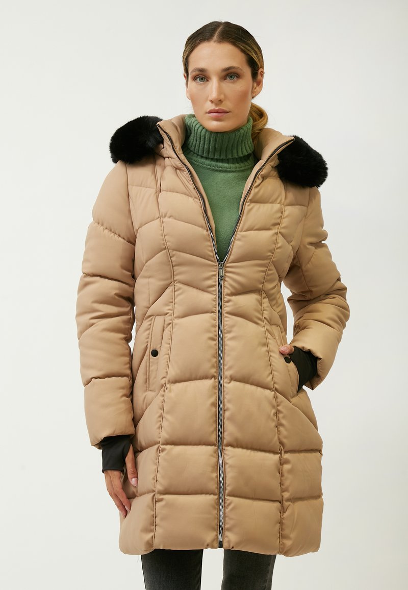 Cappotto imbottito beige a quadretti con un accento di pelliccia nera sul cappuccio, chiusura zip frontale e tasche laterali, indossato sopra un dolcevita verde.