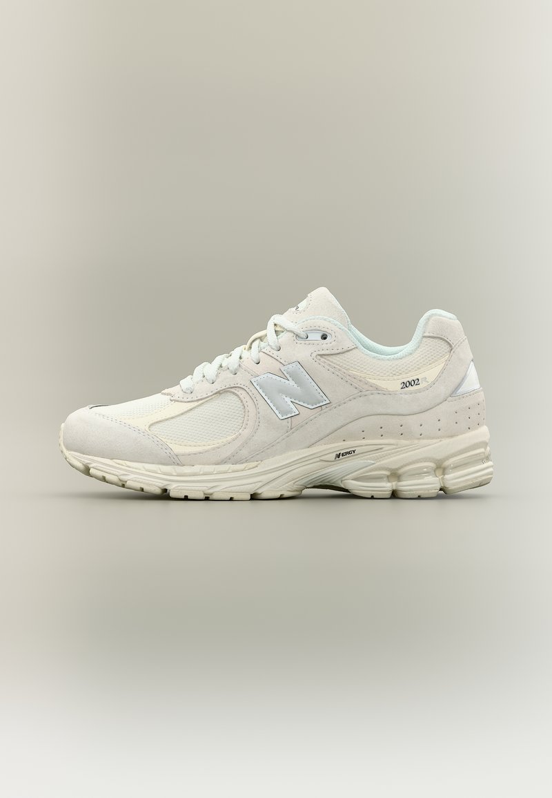 Μπεζ παπούτσι τρεξίματος New Balance 2002 με επάνω μέρος από διχτυωτό ύφασμα και σουέντ, ανακλαστικό λογότυπο "N" και αποσβεσμένη σόλα, εμφανίζεται από το πλάι σε ουδέτερο φόντο.